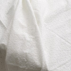 May include: Close-up of white eyelet fabric with a delicate, textured pattern. The fabric is draped, showing folds and shadows that highlight the intricate circular designs. This textile is suitable for clothing and home decor.