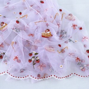 Puede incluir: Una tela transparente, de color lila claro, adornada con bordados de fresas, lazos y porciones de pastel. La tela tiene un borde festoneado con corazones rojos y la palabra "strawberry".