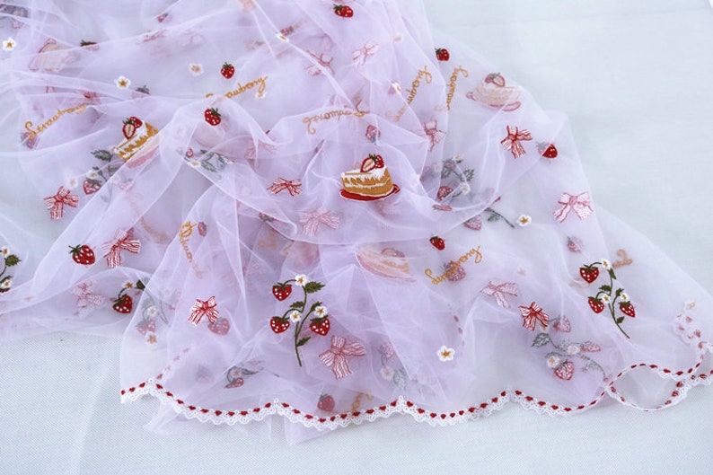 Puede incluir: Una tela transparente, de color lila claro, adornada con bordados de fresas, lazos y porciones de pastel. La tela tiene un borde festoneado con corazones rojos y la palabra "strawberry".