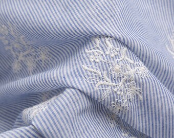 May include: Close-up of a blue and white striped fabric with white embroidered flowers. The fabric is soft and textured.