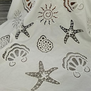 Puede incluir: Tela blanca con diseños recortados de estrellas de mar, conchas marinas y soles. La tela tiene una apariencia delicada y de encaje, con patrones intrincados que crean un tema de playa o náutico. Las formas recortadas están espaciadas uniformemente.