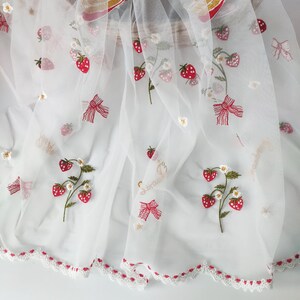 Puede incluir: Tela blanca transparente con bordados de fresas rojas, lazos y detalles florales. La tela tiene un borde festoneado con ribete rojo y la palabra "strawberry" en escritura dorada. Un dise&ntilde;o delicado y caprichoso.