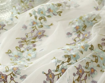 Puede incluir: Tela blanca transparente con bordados florales. El bordado presenta flores de color azul claro, morado y amarillo con tallos y hojas verdes. La tela está drapeada, creando pliegues y sombras suaves.