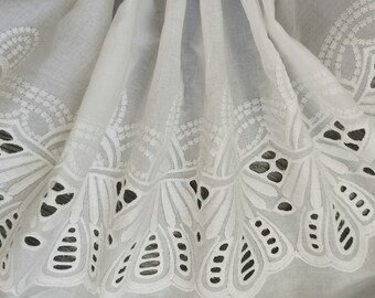 May include: White cotton fabric with a delicate, embroidered lace trim. The lace features a repeating pattern of stylized leaves and circles.