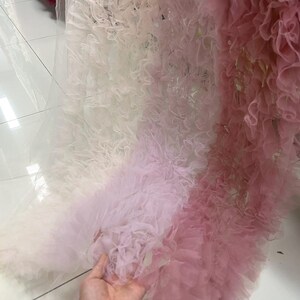May include: A close-up of layered fabric in shades of white, light pink, and deep pink. The fabric has a ruffled texture, creating a soft, voluminous appearance. The material appears to be tulle or a similar lightweight textile, suitable for dresses or decorative projects.