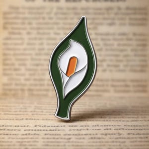May include: A green, white, and orange enamel pin in the shape of a calla lily. The pin has a silver-colored outline and is set against a blurred background of old newspaper text.