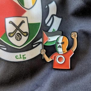 May include: An enamel pin depicting a figure in an orange and green mask, holding a Palestinian flag and raising a fist. The pin is set against a dark fabric background with a crest and the letters "c.l.g."