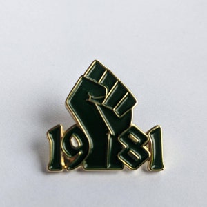Puede incluir: Un pin de color dorado con un puño levantado verde y el año "1981" en letras doradas.