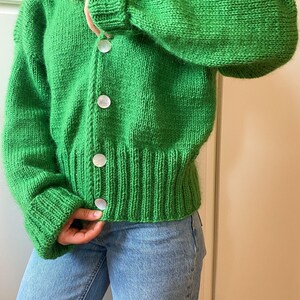 Könnte beinhalten: Ein leuchtend grüner, grob gestrickter Cardigan mit großen, perlmuttartigen Knöpfen. Der Pullover hat einen gerippten Saum und Bündchen, und die Ärmel sind leicht aufgebauscht. Der Cardigan wird mit hellblauen Jeans getragen.