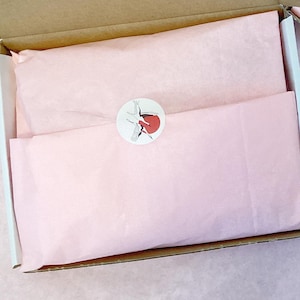 Könnte beinhalten: Eine offene Pappschachtel mit rosa Seidenpapier, einem runden Aufkleber mit rotem Design und einem Etikett mit japanischem Text. Die Schachtel steht auf einer rosa Oberfläche.