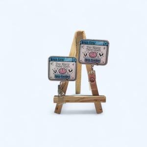 May include: Two small, rectangular pins with a computer-like design, each displaying the text "Brain Error," "Too Many Tabs Open," and "Open Another." The pins are light blue, white, and pink, and are clipped to a small wooden easel.