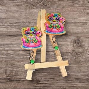 Peut inclure: Deux enrouleurs de badge colorés avec une grenouille de dessin animé portant des lunettes de soleil et une couronne de fleurs, flottant sur un flotteur de flamant rose. Les enrouleurs ont une couverture en plastique transparent et un cordon rétractable avec un bouton vert.