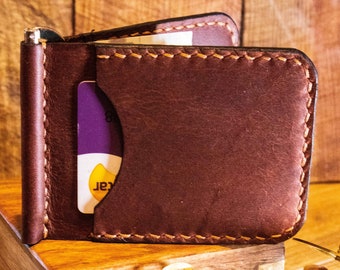 May include: A brown leather wallet with a money clip and a card slot. The wallet is closed and sitting on a wooden box.