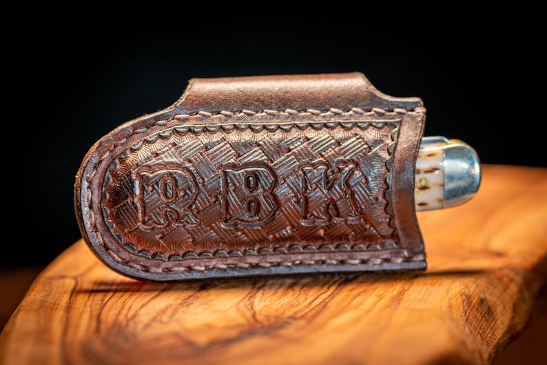 K&ouml;nnte beinhalten: Eine braune Leder-Messerscheide mit den Initialen "RBK" auf der Vorderseite. Die Scheide hat ein gewebtes Muster und ist an den Kanten gen&auml;ht. Ein silbernes und knochenfarbenes Messer ist teilweise sichtbar.