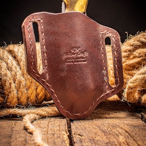 May include: A brown leather knife sheath with a stitched design and the text "Leather Craft 100% Leather 100% Handmade".
