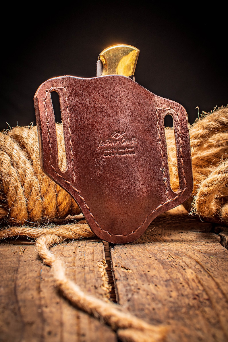 May include: A brown leather knife sheath with a stitched design and the text "Leather Craft 100% Leather 100% Handmade".