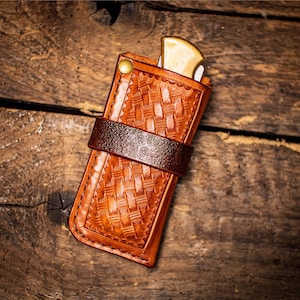 May include: A brown leather knife sheath with a woven pattern and a brown leather strap. The sheath is designed to hold a pocket knife.