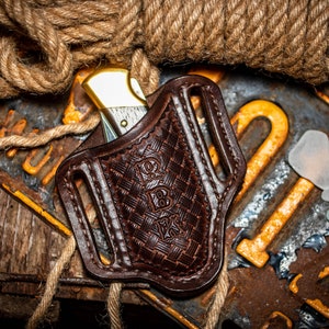 May include: A brown leather knife sheath with a woven pattern and a gold-colored pocket knife inside. The sheath has a belt loop and is sitting on a wooden surface with a rope and a rusty metal plate.