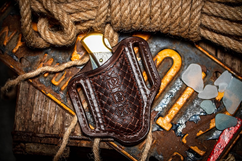 May include: A brown leather knife sheath with a woven pattern and a gold-colored pocket knife inside. The sheath has a belt loop and is sitting on a wooden surface with a rope and a rusty metal plate.