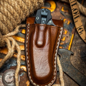 May include: A brown leather knife sheath with a white stitch detail. The sheath is empty, but a black folding knife is visible in the background.