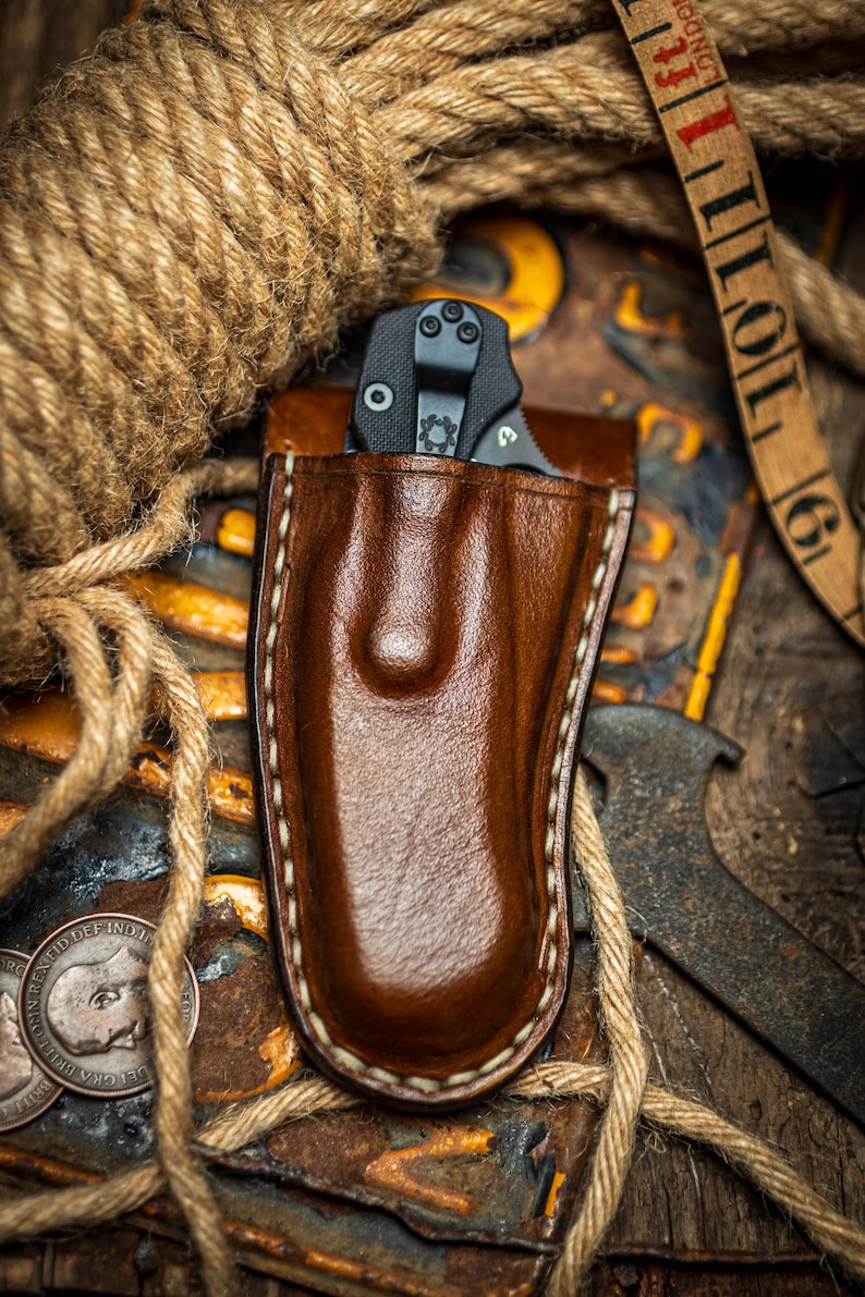 May include: A brown leather knife sheath with a white stitch detail. The sheath is empty, but a black folding knife is visible in the background.