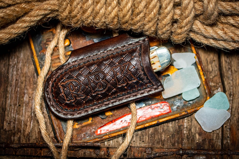 K&ouml;nnte beinhalten: Eine braune Leder-Messerscheide mit dem gepr&auml;gten Wort "ROAM". Sie enth&auml;lt ein kleines Taschenmesser mit silberner Klinge und cremefarbenem Griff. Das Bild ist vor einem rustikalen Hintergrund mit Seil und Meerglas platziert.