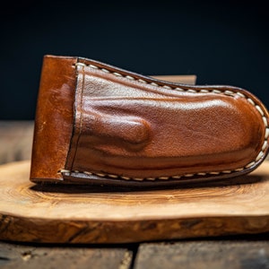 May include: A brown leather knife sheath with white stitching. The sheath is sitting on a wooden surface.