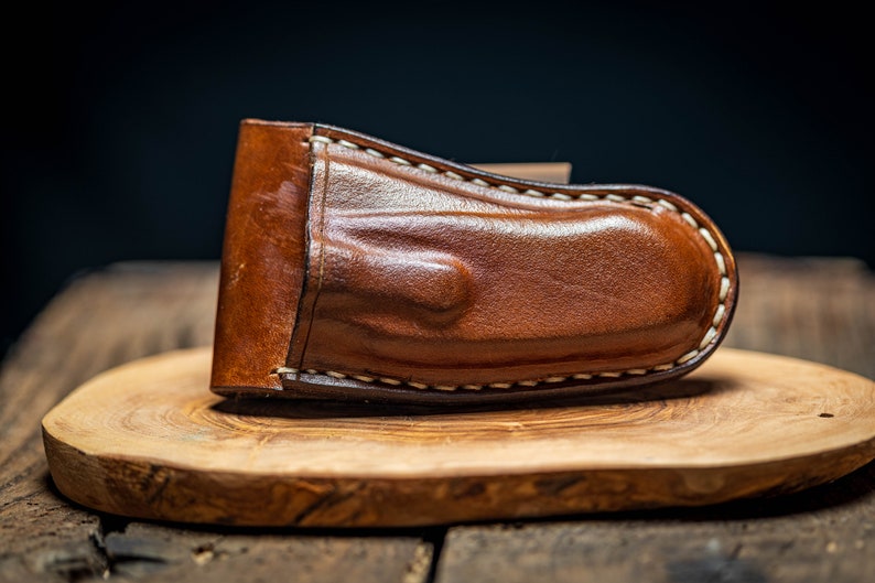 May include: A brown leather knife sheath with white stitching. The sheath is sitting on a wooden surface.