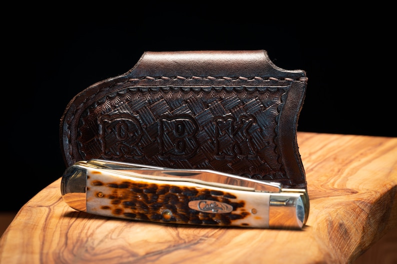 K&ouml;nnte beinhalten: Ein geschlossenes Taschenmesser mit braunem und beigem Griff liegt in einer dunkelbraunen Lederscheide. Die Scheide hat ein Webmuster und die Initialen "RBW" sind eingepr&auml;gt. Das Messer hat eine silberne Klinge und das Wort "Case" ist sichtbar.