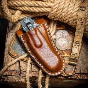 May include: A brown leather knife sheath with a black folding knife inside. The sheath has white stitching around the edges. The knife sheath is sitting on a wooden surface with rope and coins.
