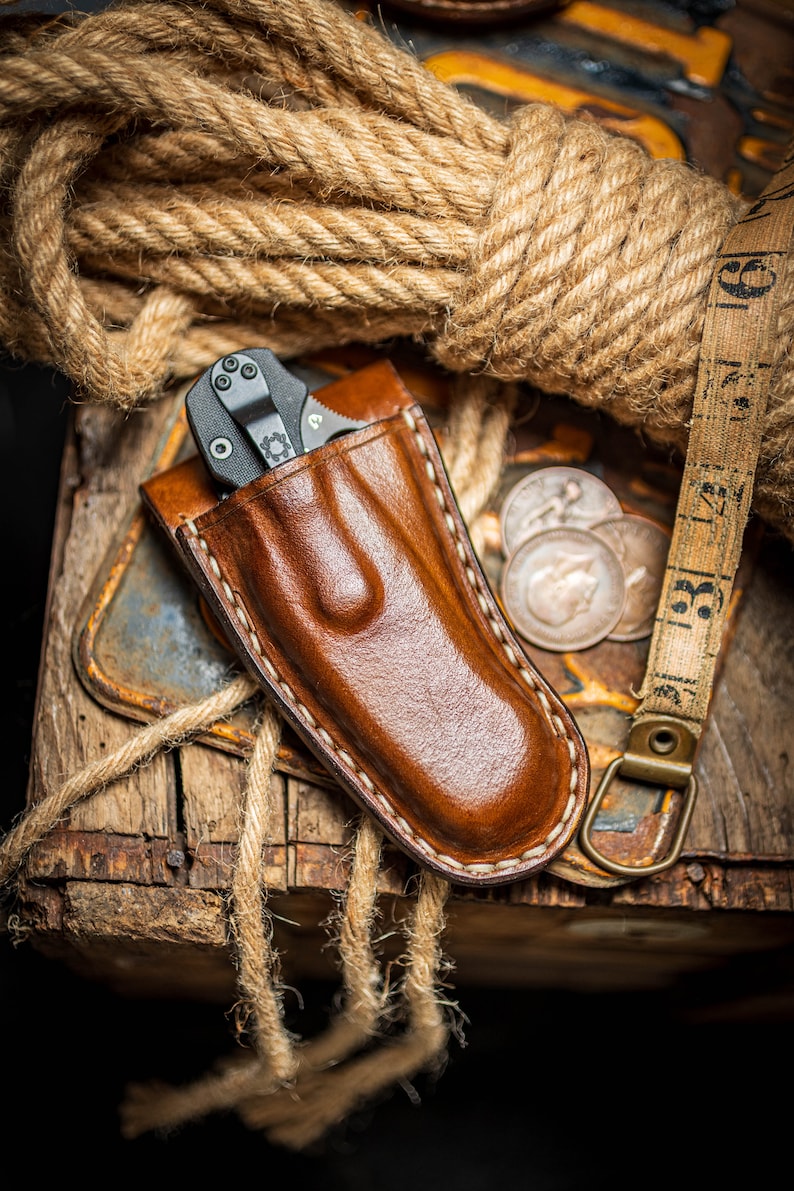 May include: A brown leather knife sheath with a black folding knife inside. The sheath has white stitching around the edges. The knife sheath is sitting on a wooden surface with rope and coins.