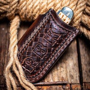 K&ouml;nnte beinhalten: Eine braune Leder-Messerscheide mit gepr&auml;gtem Design, die ein teilweise sichtbares Taschenmesser enth&auml;lt. Die Scheide liegt auf einer Holzoberfl&auml;che mit einem aufgerollten Seil im Hintergrund. Das Messer hat eine silberne Klinge und einen cremefarbenen Griff.