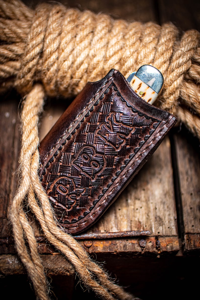 K&ouml;nnte beinhalten: Eine braune Leder-Messerscheide mit gepr&auml;gtem Design, die ein teilweise sichtbares Taschenmesser enth&auml;lt. Die Scheide liegt auf einer Holzoberfl&auml;che mit einem aufgerollten Seil im Hintergrund. Das Messer hat eine silberne Klinge und einen cremefarbenen Griff.