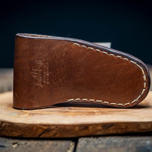 May include: A brown leather knife sheath with white stitching. The sheath has the text "Leather Goods" embossed on the front.