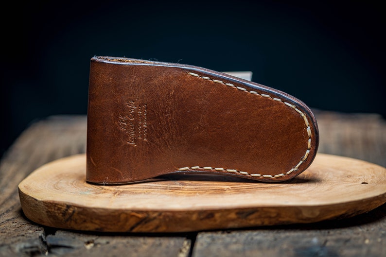 May include: A brown leather knife sheath with white stitching. The sheath has the text "Leather Goods" embossed on the front.