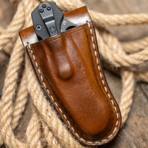 May include: A brown leather knife sheath with white stitching. A black pocket knife is inside the sheath.