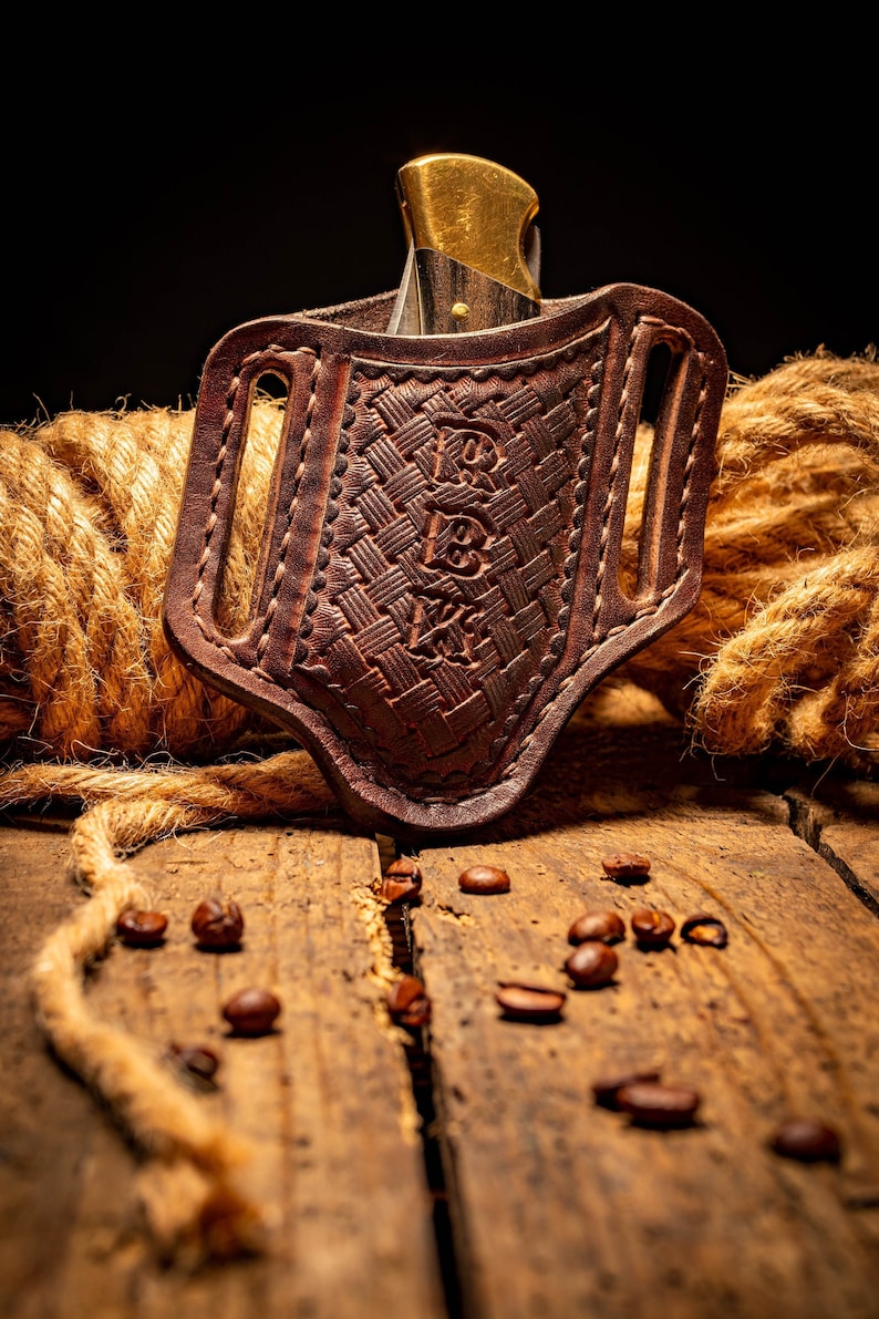 May include: A brown leather knife sheath with a woven pattern and a gold-colored knife inside. The sheath is sitting on a wooden surface with coffee beans scattered around it.