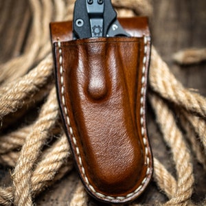May include: A brown leather knife sheath with white stitching. A black folding knife is inside the sheath.