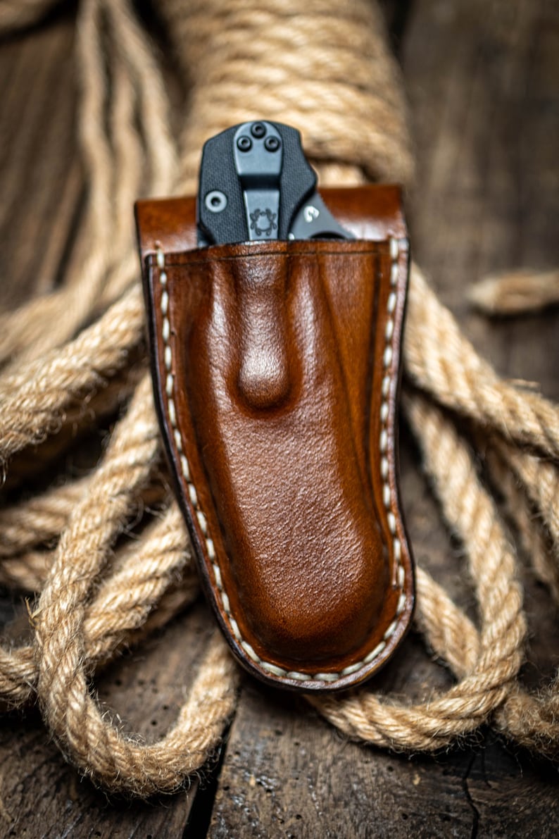 May include: A brown leather knife sheath with white stitching. A black folding knife is inside the sheath.