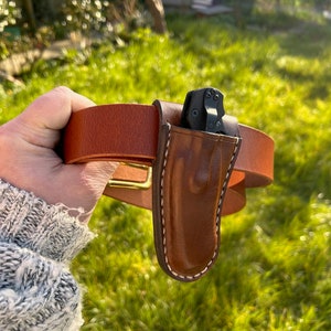 May include: A brown leather belt with a brass buckle and a brown leather sheath for a small black folding knife.