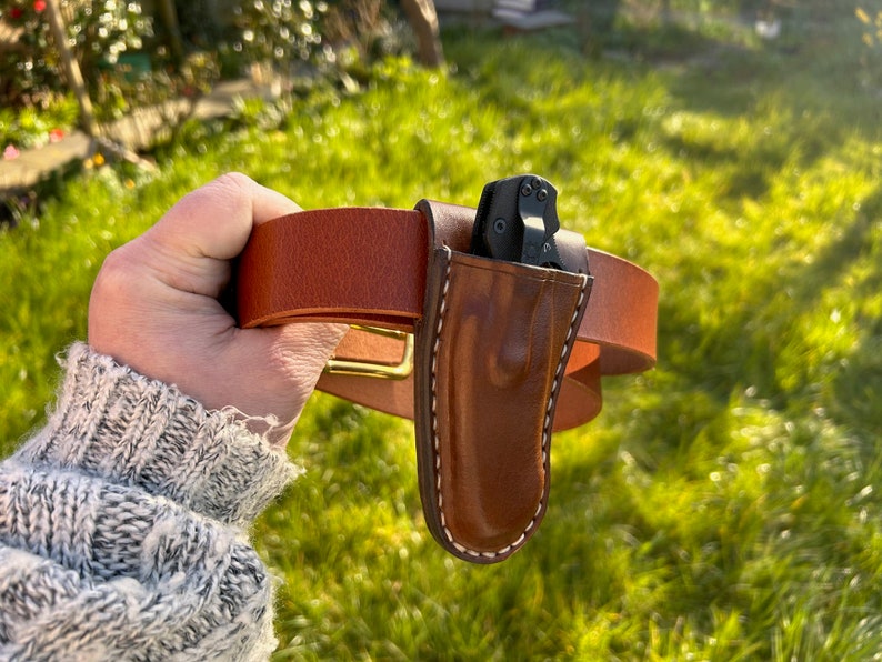May include: A brown leather belt with a brass buckle and a brown leather sheath for a small black folding knife.