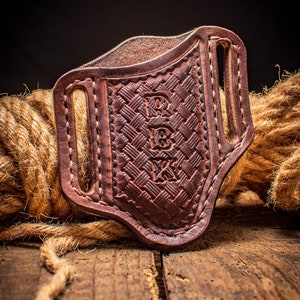May include: A brown leather knife sheath with a woven pattern and the initials "R.B.V." embossed on the front. The sheath has a belt loop and is resting on a wooden surface.