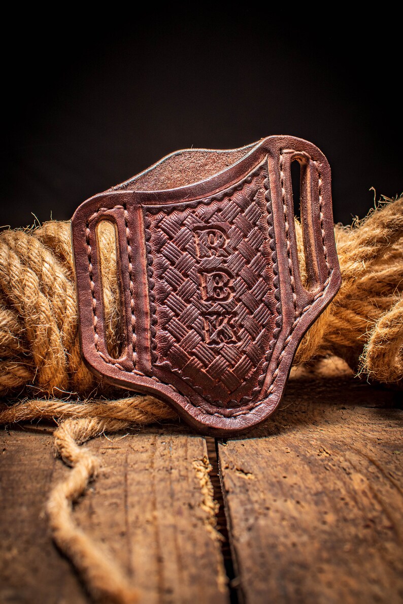 May include: A brown leather knife sheath with a woven pattern and the initials "R.B.V." embossed on the front. The sheath has a belt loop and is resting on a wooden surface.
