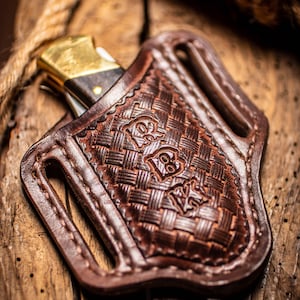 May include: A brown leather knife sheath with a woven pattern and a stitched border. The sheath has a belt loop and is designed to hold a folding knife.