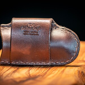 K&ouml;nnte beinhalten: Ein braunes Lederportemonnaie mit geschwungenem Design, mit dem gepr&auml;gten Text "Leather Craft 100% Leather 100% Handmade". Das Portemonnaie ist mit einem Kontrastfaden gen&auml;ht und liegt auf einer Holzoberfl&auml;che.