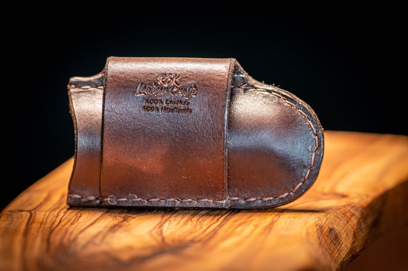 K&ouml;nnte beinhalten: Ein braunes Lederportemonnaie mit geschwungenem Design, mit dem gepr&auml;gten Text "Leather Craft 100% Leather 100% Handmade". Das Portemonnaie ist mit einem Kontrastfaden gen&auml;ht und liegt auf einer Holzoberfl&auml;che.
