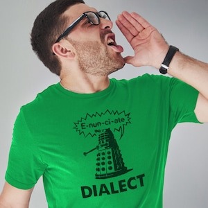 Può includere: Una maglietta verde con una grafica in bianco e nero di un Dalek di Doctor Who. Il Dalek dice "E-nun-ci-ate" in un fumetto. Il testo "Dialect" è sotto il Dalek.