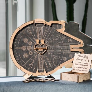 May include: A wooden guest book shaped like the Millennium Falcon from Star Wars. The guest book has a heart-shaped opening with the couple's names and wedding date engraved on it. The sign reads "Guest Book, Please sign a and drop it into the frame so we will never forget our loved ones who came."