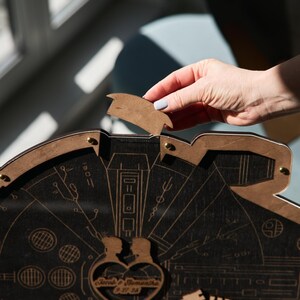May include: A wooden Millennium Falcon-shaped box with a heart-shaped cutout in the center. The box is open and a hand is reaching in to remove a small wooden piece. The heart-shaped cutout has the words "Love & Samantha" engraved on it.