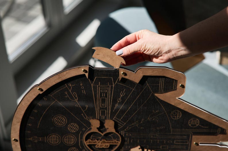 May include: A wooden Millennium Falcon-shaped box with a heart-shaped cutout in the center. The box is open and a hand is reaching in to remove a small wooden piece. The heart-shaped cutout has the words "Love & Samantha" engraved on it.